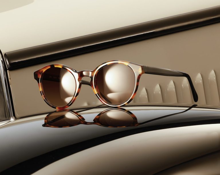 Designerbrille auf Kotflügel Runde Designerbrille mit blauem Rahmen und gemusterten Bügeln auf der Motorhaube eines Oldtimers neben einem Scheinwerfer.